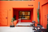 A modern outdoor café with a bold orange facade, featuring a seating area with black metal tables and chairs. There are various potted plants, including tall cacti and succulents, adding a touch of greenery. The café name, 'PlusCaffe,' is displayed above a large window.