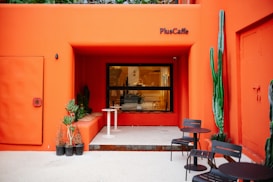 A modern outdoor café with a bold orange facade, featuring a seating area with black metal tables and chairs. There are various potted plants, including tall cacti and succulents, adding a touch of greenery. The café name, 'PlusCaffe,' is displayed above a large window.
