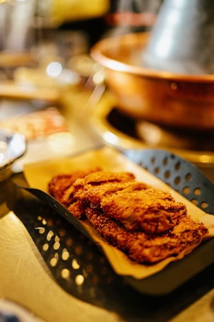A serving tray containing several pieces of crispy, golden-brown fried food, possibly chicken or another breaded dish, is placed on a surface. The food is on a sheet of parchment paper inside a perforated metal basket with a rustic, warm aesthetic. In the blurred background, there is a copper-colored pot.