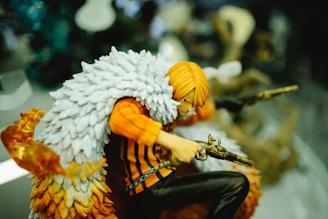 A detailed figurine of an animated character wielding dual firearms is depicted. The character has bright orange hair and wears a striking white feathered cape over an orange and black striped outfit. The figure is in a crouched, dynamic pose with one gun pointed forward and the other aimed to the side. The vibrant color scheme and intricate detailing add a lively and action-oriented feel to the figure.