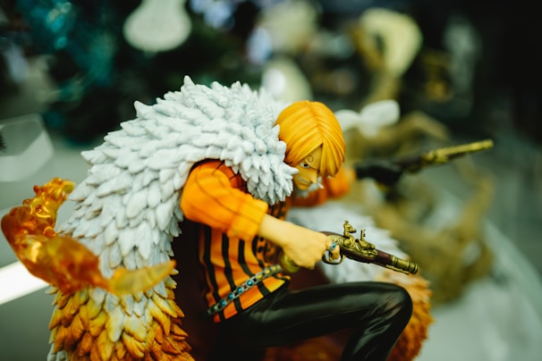 A detailed figurine of an animated character wielding dual firearms is depicted. The character has bright orange hair and wears a striking white feathered cape over an orange and black striped outfit. The figure is in a crouched, dynamic pose with one gun pointed forward and the other aimed to the side. The vibrant color scheme and intricate detailing add a lively and action-oriented feel to the figure.
