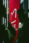 A tall, red vertical banner with white Chinese characters hangs indoors, partially obscured by large dark green leaves. Soft lighting is visible in the background, with hints of a modern interior setting.