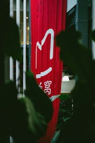 A tall, red vertical banner with white Chinese characters hangs indoors, partially obscured by large dark green leaves. Soft lighting is visible in the background, with hints of a modern interior setting.