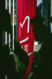 A tall, red vertical banner with white Chinese characters hangs indoors, partially obscured by large dark green leaves. Soft lighting is visible in the background, with hints of a modern interior setting.