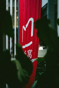 A tall, red vertical banner with white Chinese characters hangs indoors, partially obscured by large dark green leaves. Soft lighting is visible in the background, with hints of a modern interior setting.