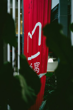 A tall, red vertical banner with white Chinese characters hangs indoors, partially obscured by large dark green leaves. Soft lighting is visible in the background, with hints of a modern interior setting.