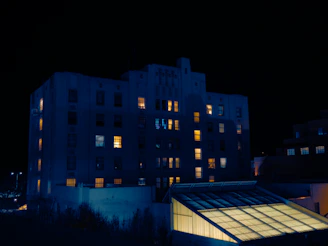 A large building with multiple stories illuminated by yellow and blue lights against a dark night sky. Several windows emit a warm yellow glow, indicating lights on inside, while others remain dark. In front of the building, a smaller structure with a glass roof is visible, lit from within.