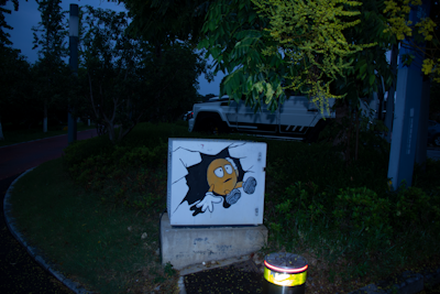 A street art piece on a utility box features a cartoonish character with a surprised expression, seemingly emerging from the wall. The surroundings are lush with green foliage, and there's a white vehicle partially visible in the background. The scene is set in low lighting, suggesting evening or nighttime.