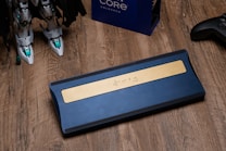 A blue mechanical keyboard with a golden accent lies on a wooden surface. To the left, there are two robotic legs, suggesting a figure or model. A gaming controller is visible on the right, and a box labeled 'Core Unlocked' is partially visible in the background.
