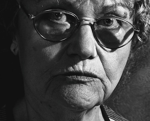 A close-up black and white photograph of an elderly person with a thoughtful or somber expression. The image focuses on their face, highlighting the texture of their skin and the reflections in their glasses. The lighting creates strong contrasts and deep shadows.