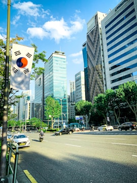 A city street flanked by tall modern skyscrapers under a bright blue sky with scattered clouds. A South Korean flag is prominently displayed on a pole beside the road. Several vehicles, including a car and a motorcycle, are visible on the street, moving in different directions. Trees line the street, adding a touch of greenery to the urban setting.