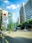 A city street flanked by tall modern skyscrapers under a bright blue sky with scattered clouds. A South Korean flag is prominently displayed on a pole beside the road. Several vehicles, including a car and a motorcycle, are visible on the street, moving in different directions. Trees line the street, adding a touch of greenery to the urban setting.