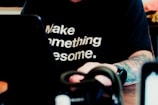 A person with a tattooed arm is wearing a black shirt with the motivational phrase 'Make something awesome.' The individual is sitting at a table, working on a laptop. The focus is on the text of the shirt, with the rest of the scene being slightly blurred.