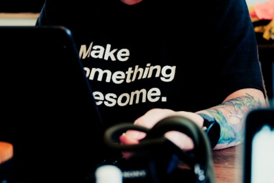 A person with a tattooed arm is wearing a black shirt with the motivational phrase 'Make something awesome.' The individual is sitting at a table, working on a laptop. The focus is on the text of the shirt, with the rest of the scene being slightly blurred.