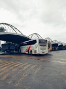 A bus station with a large white bus prominently displayed. The bus has bold red and black graphics on its side and is parked on a paved area with yellow lines marking the ground. The station has a series of large, curved metal awnings overhead, with advertisements visible in the background. The sky is overcast, giving a dim lighting to the scene.