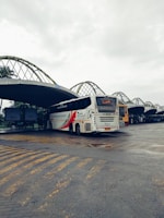 A bus station with a large white bus prominently displayed. The bus has bold red and black graphics on its side and is parked on a paved area with yellow lines marking the ground. The station has a series of large, curved metal awnings overhead, with advertisements visible in the background. The sky is overcast, giving a dim lighting to the scene.