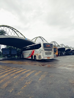 A bus station with a large white bus prominently displayed. The bus has bold red and black graphics on its side and is parked on a paved area with yellow lines marking the ground. The station has a series of large, curved metal awnings overhead, with advertisements visible in the background. The sky is overcast, giving a dim lighting to the scene.