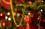 A festive Christmas tree adorned with a small plaid gift box, decorative tassel, and golden bead garlands, set against a background of glowing bokeh lights.