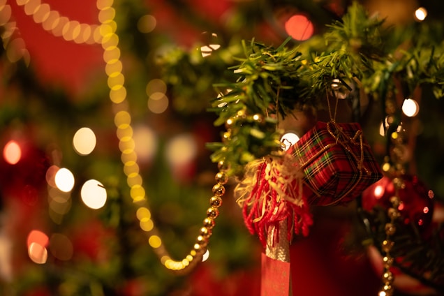 A festive Christmas tree adorned with a small plaid gift box, decorative tassel, and golden bead garlands, set against a background of glowing bokeh lights.