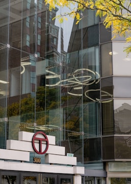 A modern glass building with reflective surfaces shows the surrounding architecture and trees. The facade features a circular logo and the number 300 prominently displayed. Yellow leaves from nearby trees add a touch of color against the mostly glass and concrete structure.