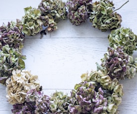 A circular wreath made of dried hydrangea flowers in various shades of green, purple, and beige is laid on a light, textured wooden surface.