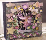 An intricate and colorful 3D card featuring celestial and festive themes. The design includes angels with yellow wings and halos, a radiant sun with a serene face, and various decorative patterns including stars, flowers, and swirling clouds. The artwork is richly adorned with sequins and ornaments, adding a layered and textured appearance.