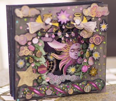 An intricate and colorful 3D card featuring celestial and festive themes. The design includes angels with yellow wings and halos, a radiant sun with a serene face, and various decorative patterns including stars, flowers, and swirling clouds. The artwork is richly adorned with sequins and ornaments, adding a layered and textured appearance.