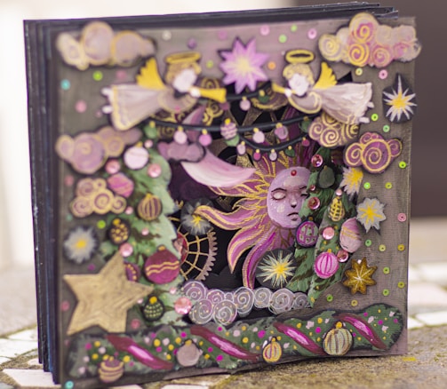 An intricate and colorful 3D card featuring celestial and festive themes. The design includes angels with yellow wings and halos, a radiant sun with a serene face, and various decorative patterns including stars, flowers, and swirling clouds. The artwork is richly adorned with sequins and ornaments, adding a layered and textured appearance.