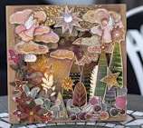 A whimsical and intricate card features an artful collage of celestial and nature elements, including stars, a crescent moon, stylized trees, clouds, floral patterns, and a serene sun face. The design is rich in details with a combination of vibrant and muted colors, enhanced by metallic and gem-like embellishments, creating a three-dimensional effect.