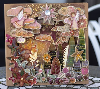A whimsical and intricate card features an artful collage of celestial and nature elements, including stars, a crescent moon, stylized trees, clouds, floral patterns, and a serene sun face. The design is rich in details with a combination of vibrant and muted colors, enhanced by metallic and gem-like embellishments, creating a three-dimensional effect.