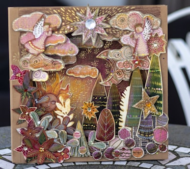 A whimsical and intricate card features an artful collage of celestial and nature elements, including stars, a crescent moon, stylized trees, clouds, floral patterns, and a serene sun face. The design is rich in details with a combination of vibrant and muted colors, enhanced by metallic and gem-like embellishments, creating a three-dimensional effect.
