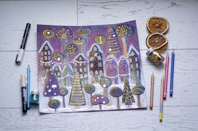 A whimsical drawing of a winter village scene with stylized houses and trees, adorned with bright dots and patterns. The artwork is surrounded by various art supplies, including colored pencils, markers, and jars of glitter. Slices of dried oranges are placed nearby on a wooden surface.