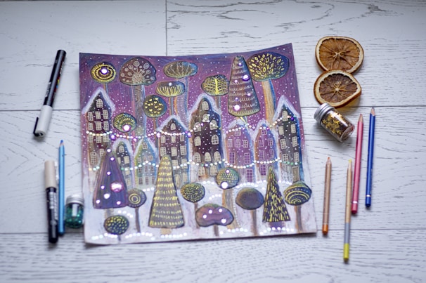 A whimsical drawing of a winter village scene with stylized houses and trees, adorned with bright dots and patterns. The artwork is surrounded by various art supplies, including colored pencils, markers, and jars of glitter. Slices of dried oranges are placed nearby on a wooden surface.