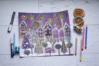 A whimsical drawing of a winter village scene with stylized houses and trees, adorned with bright dots and patterns. The artwork is surrounded by various art supplies, including colored pencils, markers, and jars of glitter. Slices of dried oranges are placed nearby on a wooden surface.