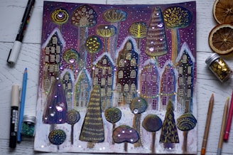 A whimsical drawing features a row of fantastical houses and unique trees adorned with intricate patterns and bright, shimmering decorations. The background includes a starry night sky with purple hues, and the foreground shows various art supplies like pencils, markers, a jar of glitter, and dried orange slices placed around the image.