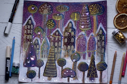 A whimsical drawing features a row of fantastical houses and unique trees adorned with intricate patterns and bright, shimmering decorations. The background includes a starry night sky with purple hues, and the foreground shows various art supplies like pencils, markers, a jar of glitter, and dried orange slices placed around the image.