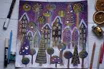 A whimsical drawing features a row of fantastical houses and unique trees adorned with intricate patterns and bright, shimmering decorations. The background includes a starry night sky with purple hues, and the foreground shows various art supplies like pencils, markers, a jar of glitter, and dried orange slices placed around the image.