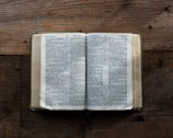 An open book lies on a textured wooden surface, displaying text in a serif font. The pages spread out evenly, revealing multiple columns of printed words. The wood shows pronounced grain patterns, adding a rustic feel to the scene.