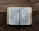 An open book lies on a textured wooden surface, displaying text in a serif font. The pages spread out evenly, revealing multiple columns of printed words. The wood shows pronounced grain patterns, adding a rustic feel to the scene.