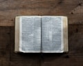 An open book lies on a textured wooden surface, displaying text in a serif font. The pages spread out evenly, revealing multiple columns of printed words. The wood shows pronounced grain patterns, adding a rustic feel to the scene.