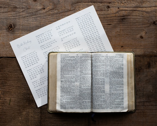 A wooden table holds an open book displaying text, likely a Bible, accompanied by a paper with a reading schedule or chart listing books and chapters.