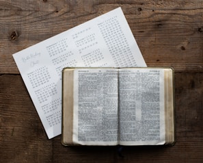 A wooden table holds an open book displaying text, likely a Bible, accompanied by a paper with a reading schedule or chart listing books and chapters.