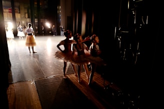 Young ballerinas in tutus are waiting backstage as a performance is underway, with warm lighting highlighting the stage and casting shadows in the wings.