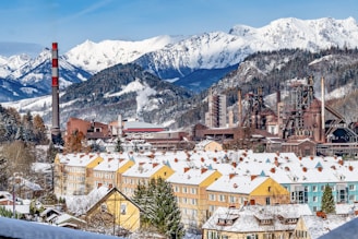 A picturesque landscape featuring snow-covered mountains in the background, with a large industrial factory complex in the foreground. The factory includes chimneys and smokestacks with visible smoke, set against a row of colorful residential buildings also topped with snow. The scene captures the juxtaposition of industrial development amidst a serene mountainous terrain.