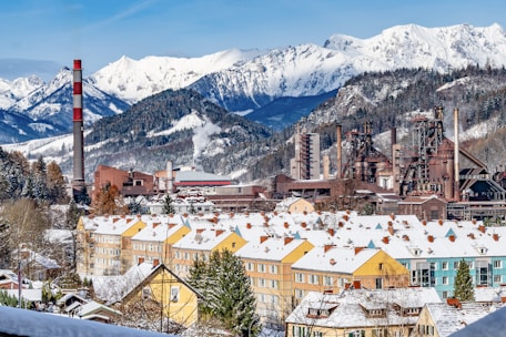 A picturesque landscape featuring snow-covered mountains in the background, with a large industrial factory complex in the foreground. The factory includes chimneys and smokestacks with visible smoke, set against a row of colorful residential buildings also topped with snow. The scene captures the juxtaposition of industrial development amidst a serene mountainous terrain.
