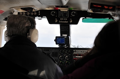 The cockpit view of a small aircraft with two people seated. The control panel is visible featuring various instruments, dials, and a small screen displaying navigational information. The person on the left is wearing headphones.