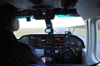 The cockpit of an aircraft with various instruments and controls is visible, indicating the airplane is in flight or preparing for flight. A pilot is seated, wearing headphones, looking towards the runway that extends straight ahead. The dashboard is equipped with numerous dials, gauges, and a digital GPS device displaying navigational data.
