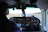 The cockpit of an aircraft with various instruments and controls is visible, indicating the airplane is in flight or preparing for flight. A pilot is seated, wearing headphones, looking towards the runway that extends straight ahead. The dashboard is equipped with numerous dials, gauges, and a digital GPS device displaying navigational data.