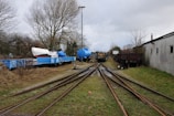 A railway track splits into two directions surrounded by grassy areas and leafless trees. On one side, there are several blue rail cars, one carrying a white covered object resembling a boat, while another has a large blue cylindrical tank. On the other side, there are some brown rail cars next to a gray building with windows and graffiti.