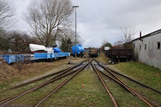 A railway track splits into two directions surrounded by grassy areas and leafless trees. On one side, there are several blue rail cars, one carrying a white covered object resembling a boat, while another has a large blue cylindrical tank. On the other side, there are some brown rail cars next to a gray building with windows and graffiti.